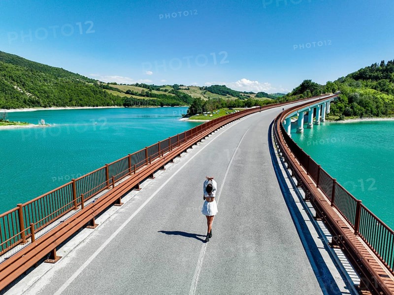 A young woman walks along a picturesque bridge overlooking a vibrant turquoise lake, surrounded by lush green hills, capturing the serene beauty of Italy's natural landscape