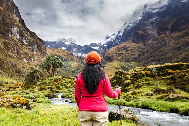 An Ecuadorian woman in a red jacket and orange hat stands by a stream, surrounded by the majestic mountains of El Altar Volcano in Ecuador, under a cloudy sky