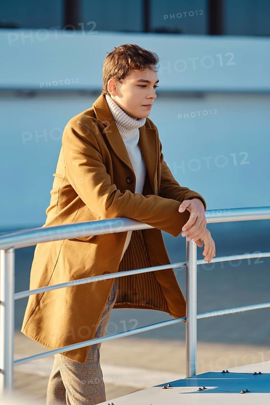 Spring, autumn outfit. Street fashion. Young man in checkered trousers, sweater and wool coat leaning to the railings