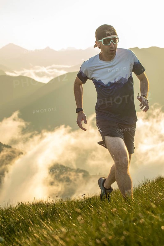 A man is trail running uphill in the early morning light, surrounded by stunning misty mountain views. He wears sunglasses, a hat, and a sporty outfit, epitomizing outdoor fitness