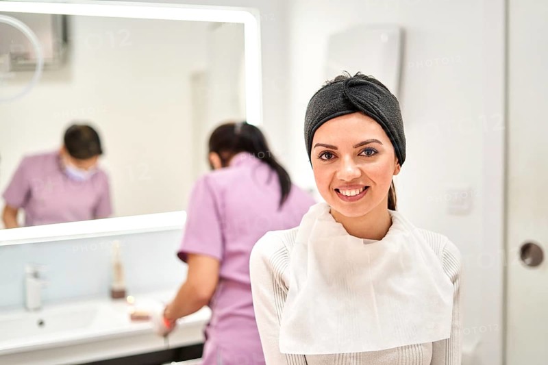 A smiling client wearing a headband and protective bib sits in a well-lit aesthetic clinic, prepared for a beauty treatment while staff work in the background, creating a welcoming atmosphere