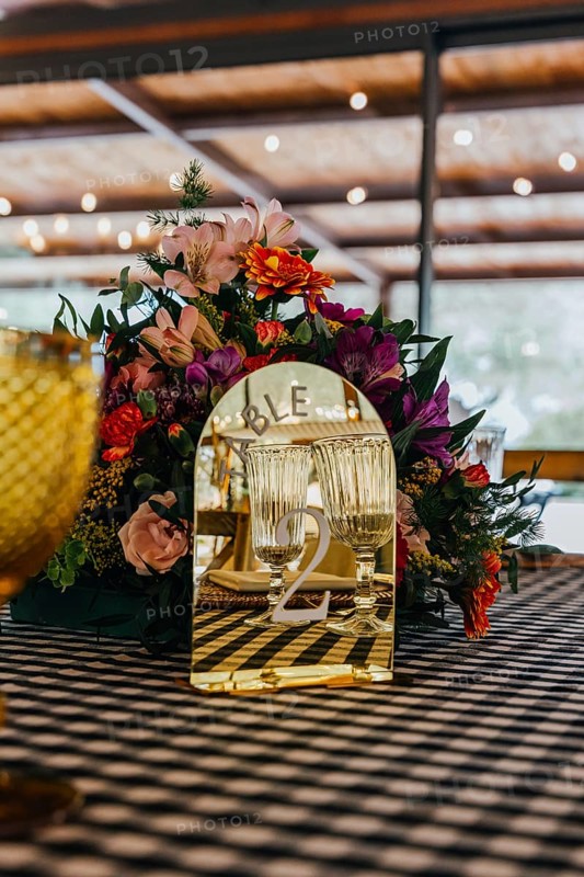 Close-up of a vibrant floral centerpiece with a golden mirror table number 2, surrounded by crystal glassware on a checkered tablecloth, creating a refined and festive atmosphere