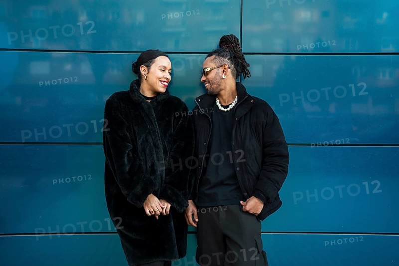 Multicultural couple wearing stylish black coats, standing against a blue wall. They are smiling warmly at each other, showcasing a moment of joy and connection
