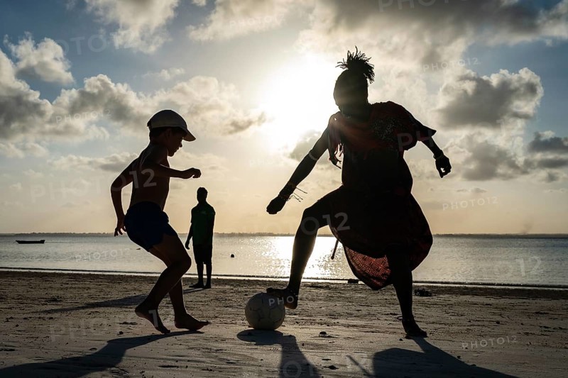 MICHAMVI KAE, ZANZIBAR - OCTOBER 14, 2025: Silhouette of a Maasai man and a young boy playing soccer on a tropical beach at sunset with the sun reflecting on water
