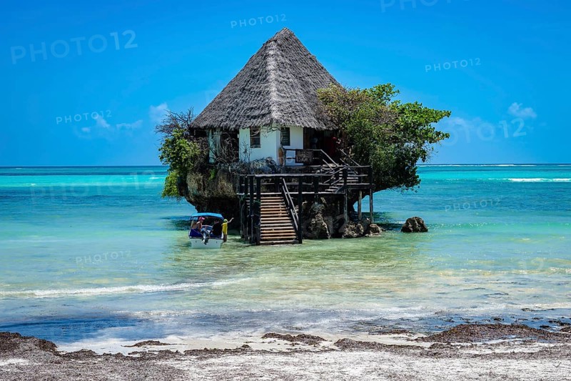 MICHAMVI PINGWE, ZANZIBAR - OCTOBER 15, 2025: The famous Rock Restaurant built on a coral outcrop in turquoise ocean water, with a small boat arriving at the wooden stairs in Zanzibar, Tanzania