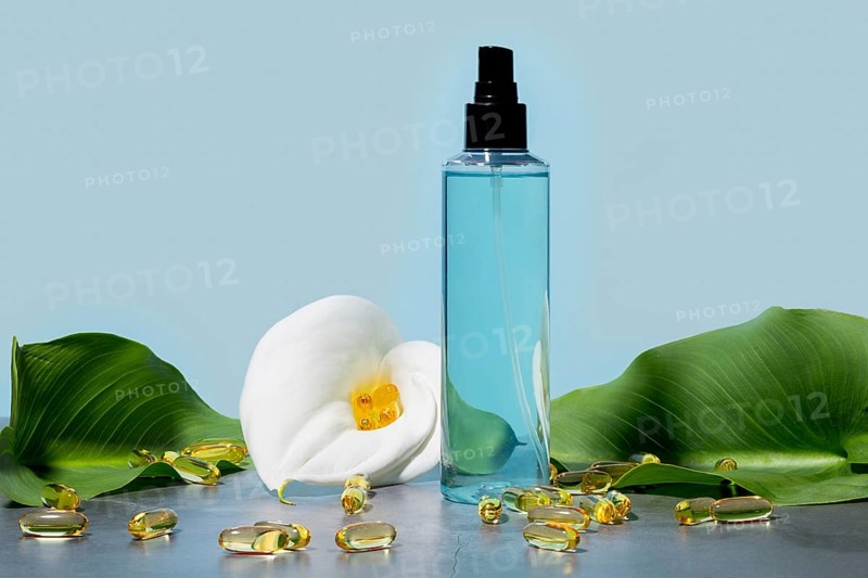 Elegant cosmetic spray clear blue bottle, paired with a fresh white calla lily and scattered omega capsules, symbolizing natural skincare solutions on a serene blue background