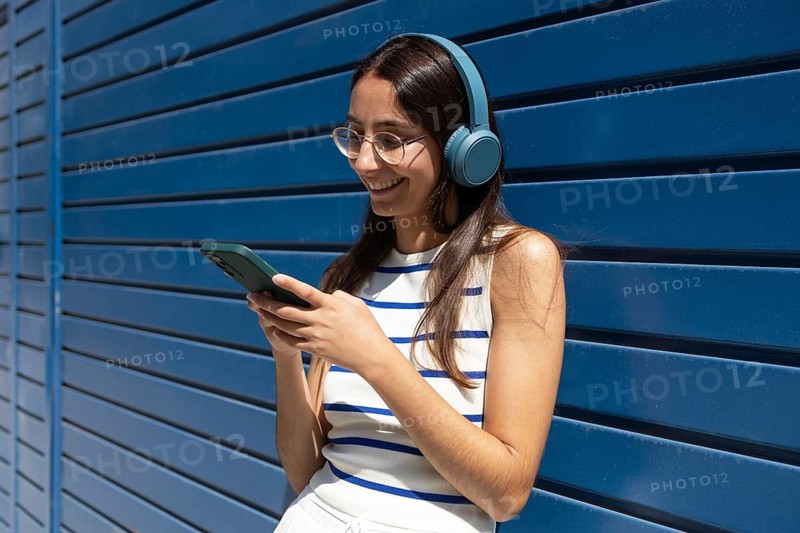 A woman in the city enjoys a sunny day, wearing headphones and using her smartphone, embodying the modern outdoor lifestyle with her relaxed and stylish demeanor