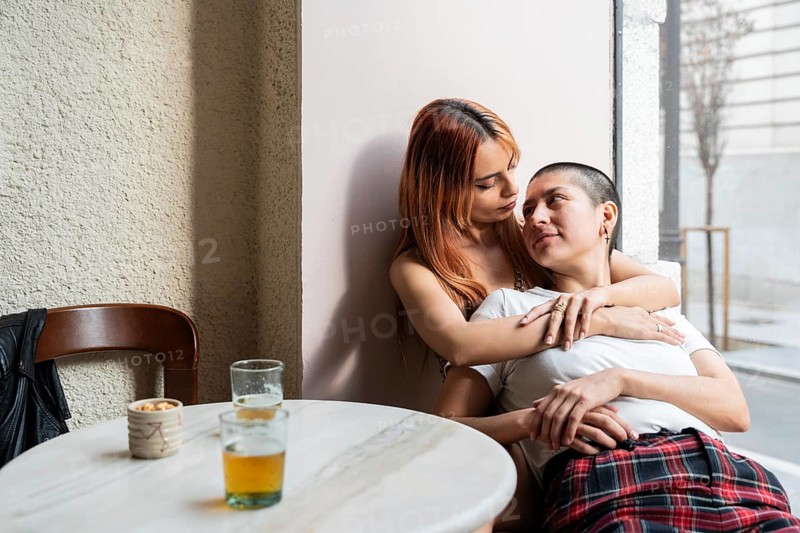 A lesbian couple enjoying a tender embrace at a café, surrounded by drinks and snacks, creating a warm and intimate atmosphere. They gaze affectionately at each other