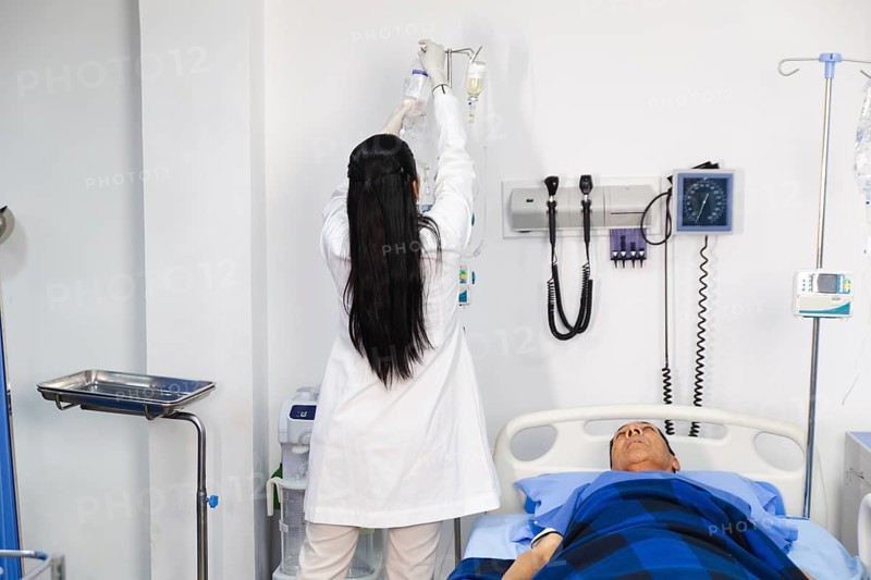 A female doctor adjusts an IV drip for an elderly patient lying in a hospital bed. The doctor, focused and attentive, ensures the patient's comfort and well-being in a clinical setting