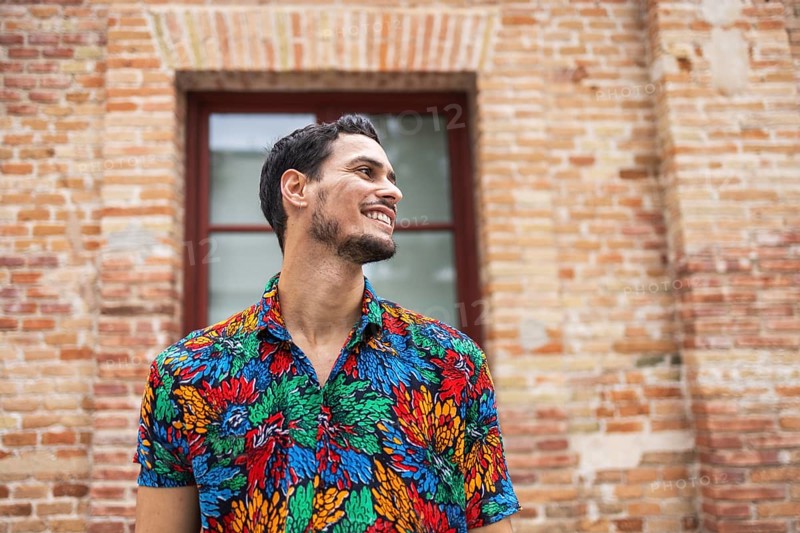 A joyful Cuban man stands against a rustic brick wall, wearing a vibrant, colorful shirt. His expression exudes happiness and warmth, capturing lively Cuban culture