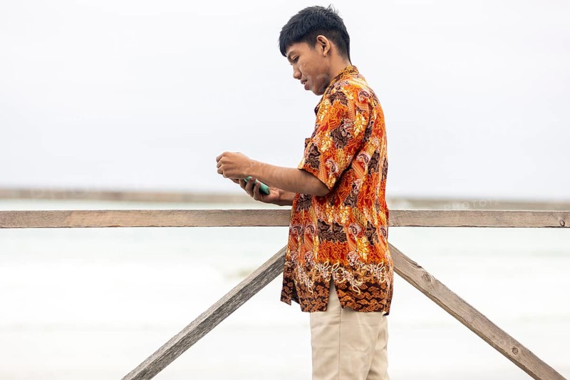 An Indonesian man stands by a wooden railing, wearing a vibrant batik shirt. The serene Australian landscape serves as a backdrop, combining cultural elements in a unique setting