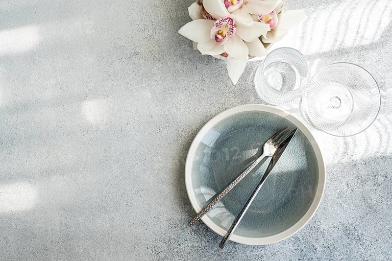Overhead view of a minimalist dining setting. A cracked gray plate with fork and knife, complemented by crystal glasses and a white orchid on a textured surface