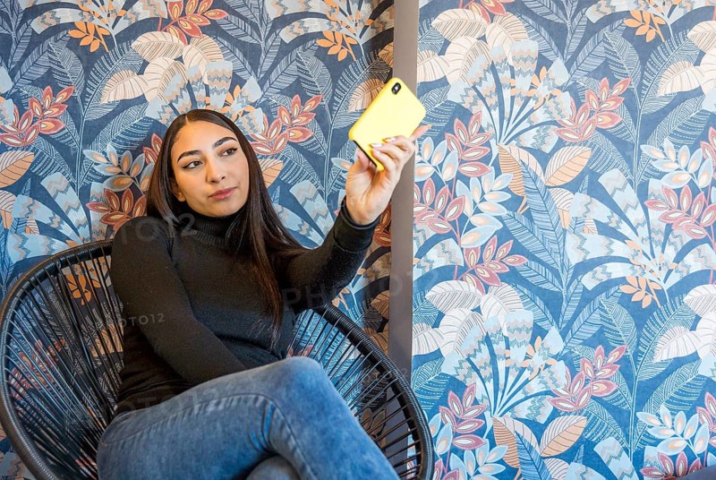 A young woman captures a selfie using her vibrant yellow mobile phone. She's seated in a stylish chair, surrounded by a beautiful floral wallpaper backdrop