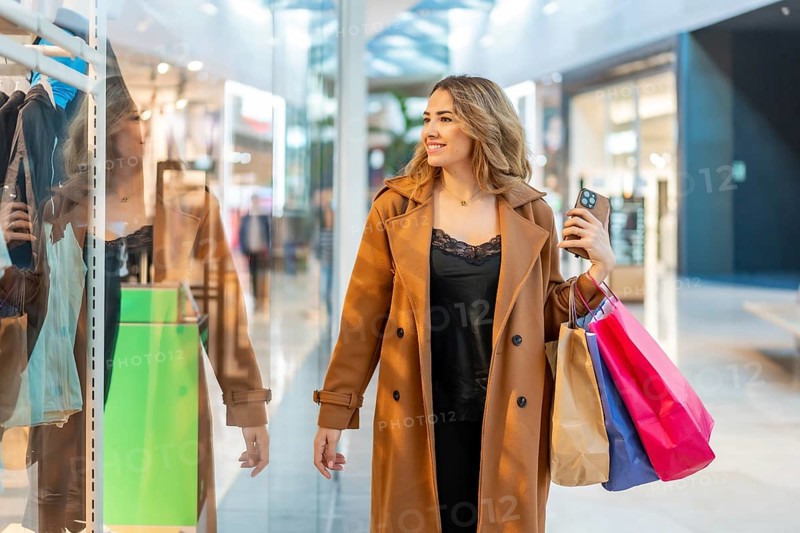 Young woman smiling while carrying multiple colorful shopping bags and a smartphone, enjoying a leisurely shopping day in a contemporary retail complex with store reflections