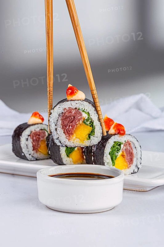 A close up view of vibrant sushi rolls filled with mango and tuna, topped with sauce. They are elegantly arranged on a white background, accompanied by a bowl of soy sauce