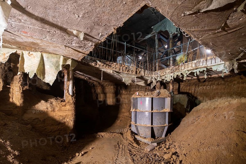 An underground scene shows a construction site in progress, with exposed soil, metal supports, and a large metal container, illuminated by natural and artificial lighting