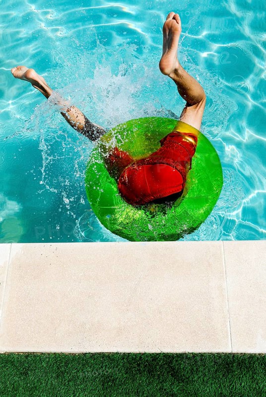 A teenage boy dives into a pool, making a splash as he lands on a green inflatable ring. He is wearing bright red shorts, creating a vibrant summer scene of fun and excitement