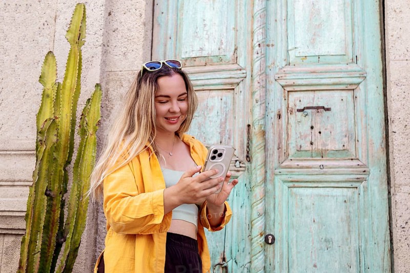 A female tourist wearing a yellow shirt and sunglasses checks her smartphone in front of a rustic turquoise door, with a cactus nearby, enjoying a moment of exploration