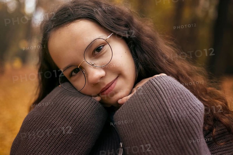A young girl with glasses, wrapped in a cozy sweater, poses against an autumn backdrop. Her smile reflects warmth and comfort in a serene outdoor setting