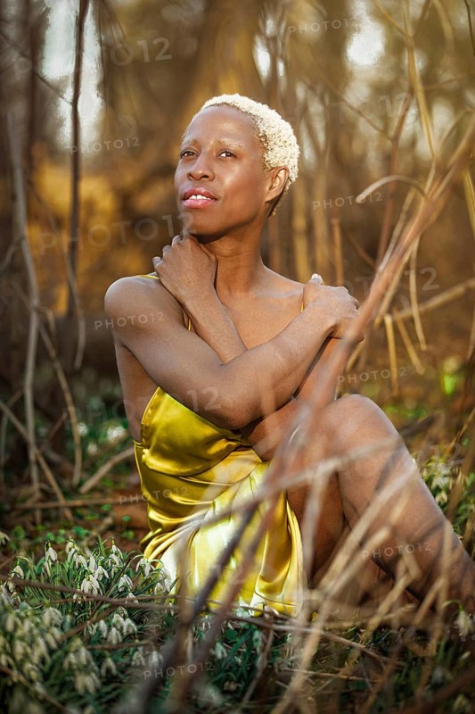 A woman in a vibrant golden dress sits thoughtfully in Tacony Creek Park. Her contemplative expression is beautifully framed by sunlit foliage and delicate blooms snowdrops