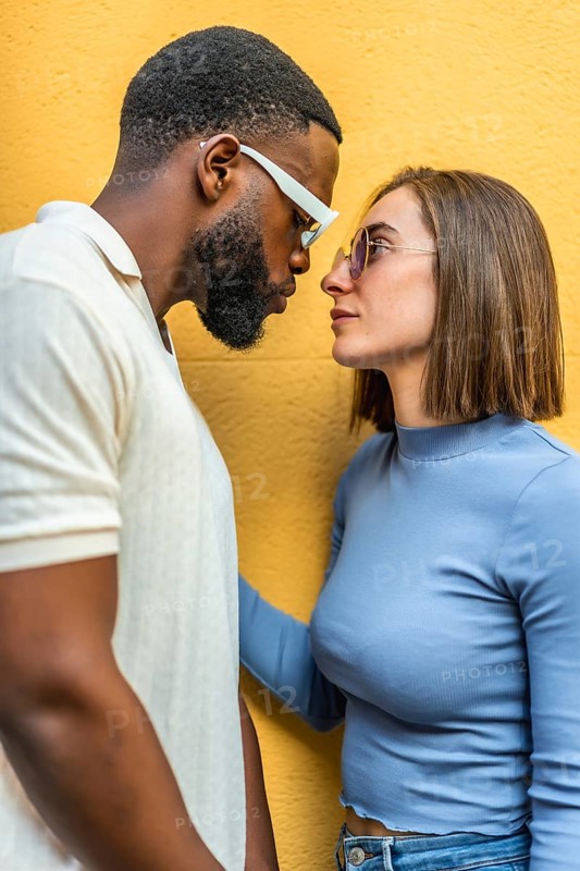 Vertical close-up side view of a cool modern couple looking at each other wearing sunglasses outdoors against yellow background