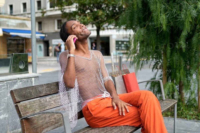 A man in vibrant fashion sits on a bench, exuding joy and pride. His mesh top and orange pants capture a spirited expression of individuality and LGBTQIA+ pride