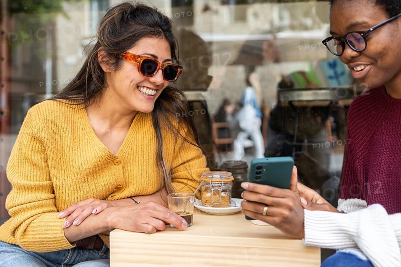 Two friends enjoy a sunny day at a cafe, sharing coffee and smiles. One wears stylish sunglasses while they browse on a smartphone together, creating a joyful atmosphere