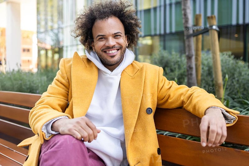 Smiling Afro man in a vibrant yellow coat enjoys a relaxed moment sitting on a bench outdoors. Modern, urban setting creates a cheerful and casual atmosphere