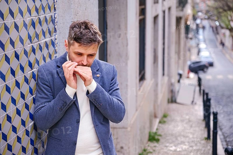 Man musician in a blue blazer and white sweater playing blues on a harmonica with eyes closed, standing outdoors in Lisbon city street, Portugal
