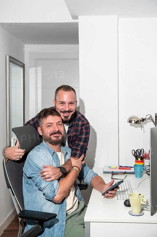 A loving gay couple is collaborating at their home office desk. One is seated, holding a smartphone, while the other lovingly embraces him from behind, both are looking at camera