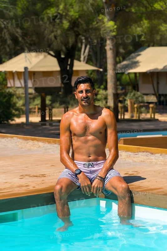 Young man is enjoying his vacation, sitting on the edge of a swimming pool with his feet in the water, at a glamping resort with canvas tents in the background, surrounded by trees