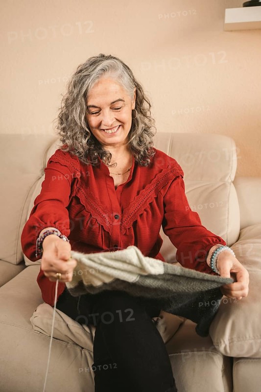 An older woman with gray hair knits contently on a beige sofa, wearing a red blouse. The cozy setting and natural light enhance the peaceful atmosphere of her hobby