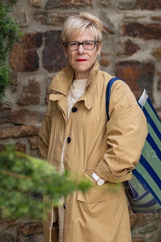 A confident mature woman in stylish autumn attire stands against a rustic stone wall. Her beige trench coat and striped bag complement the natural outdoor setting