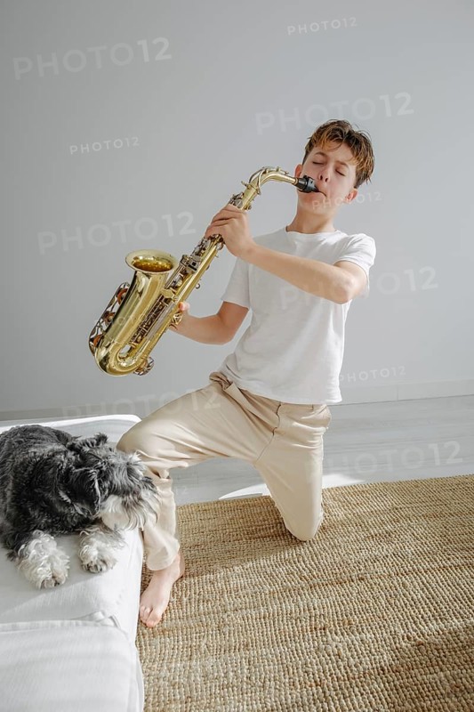 A young boy in a bright room plays a saxophone while kneeling on a carpet. His dog rests beside him, creating a harmonious and serene atmosphere