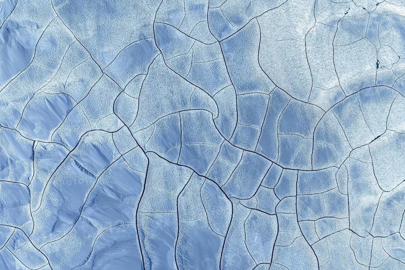 Abstract aerial image of a frozen landscape featuring intricate patterns and cracks. The blue and white tones create a mesmerizing visual effect, highlighting nature's artistry