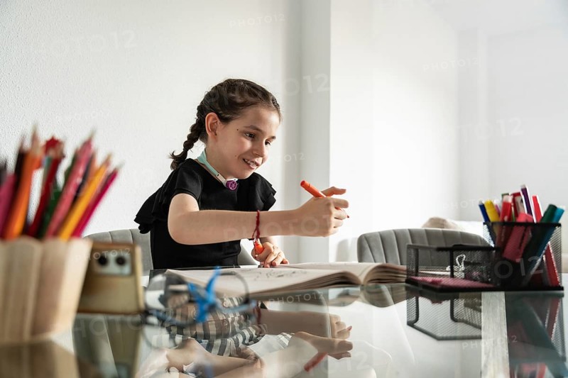 A young disabled girl with a tracheostomy happily draws at a table, surrounded by colorful pencils, in a bright, cheerful setting, showcasing her creativity and enthusiasm
