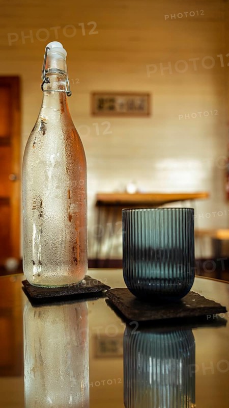 A chilled glass bottle and a textured tumbler sit on square coasters atop a reflective table. Warm, inviting cafe ambiance with soft focus in the background