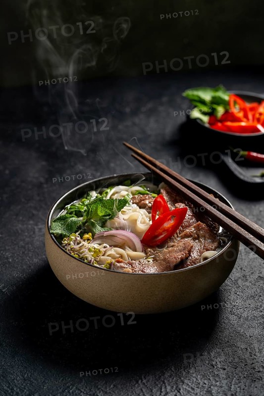 A steaming bowl of Vietnamese Beef Pho Noodle Soup (Pho Bo), garnished with slices of beef, fresh herbs, onions, and a slice of red chili, artfully presented in a dark, textured backdrop