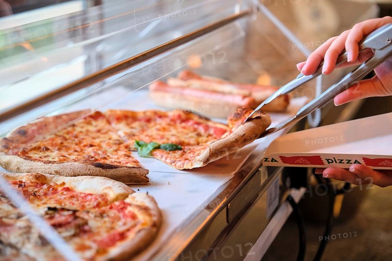 Metal tongs grabbing slice of margherita pizza from display case in pizzeria, ready to be served in cardboard box