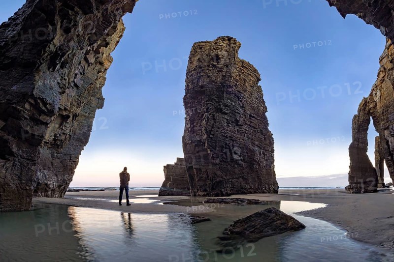 A person stands at the base of towering coastal rock formations, surrounded by reflective water from the receding tide. The scene captures the serene beauty at sunrise
