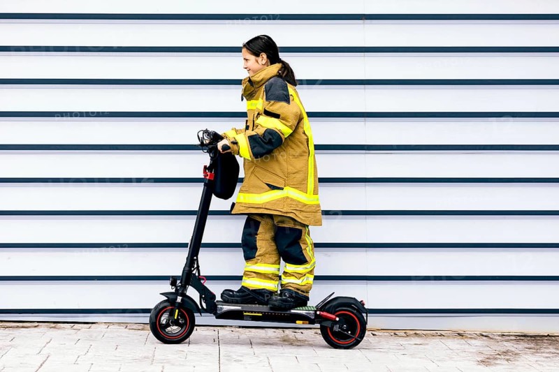 A young girl dressed in a firefighter suit rides an electric scooter in front of a striped wall, showcasing a blend of playful imagination and professional aspiration