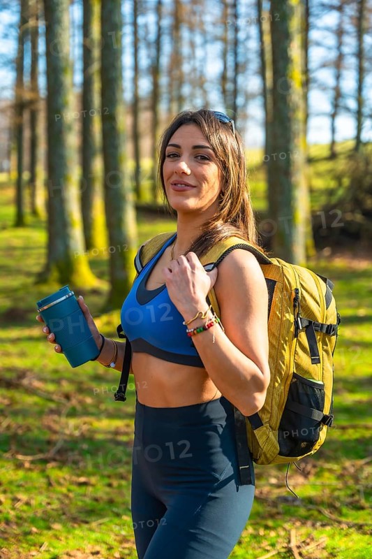 Young hiker woman walking in a forest holding a reusable coffee cup, wearing sportswear and backpack, enjoying a sunny day