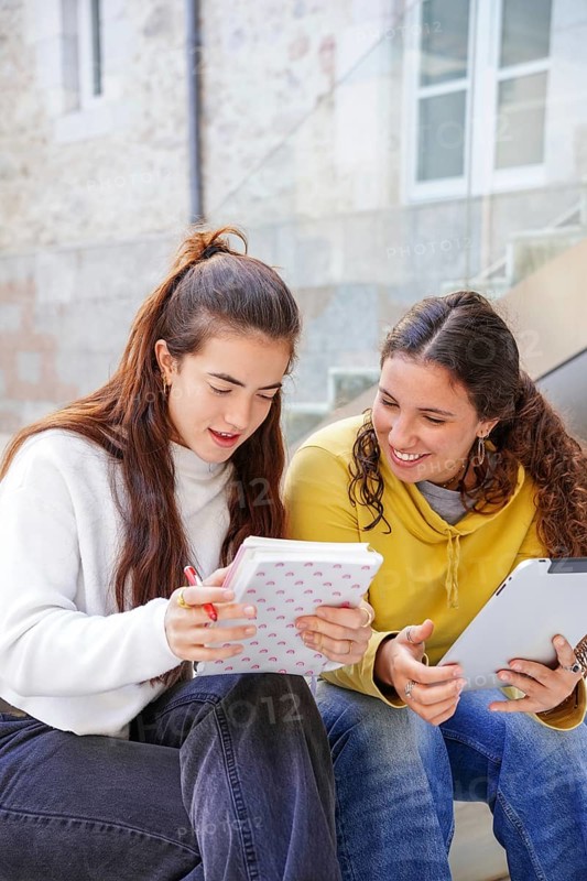 Two students sit on stairs, engaged in study and discussion One takes notes in a notebook, while the other uses a tablet Both seem focused and collaborative