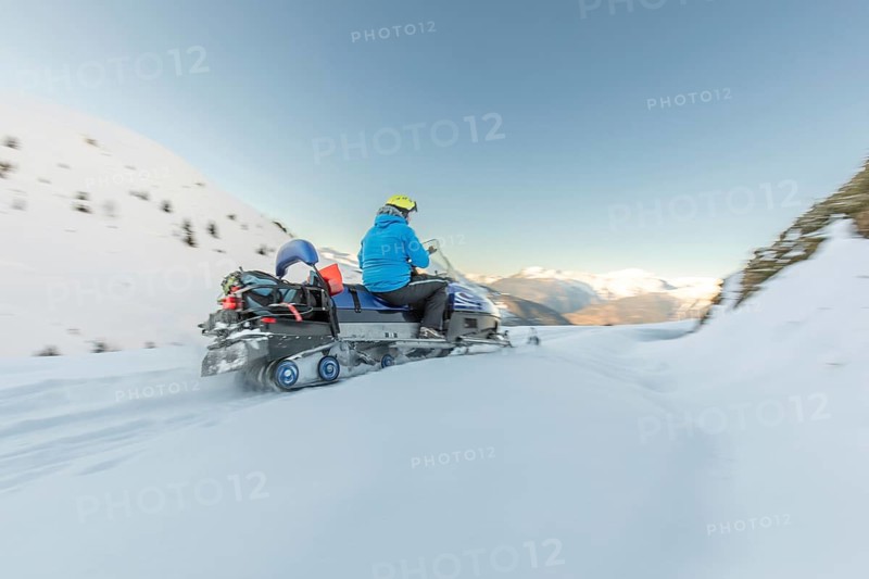 A snowmobile rider in a bright blue jacket navigates through a vast, snowy landscape on a clear winter day. Majestic snow-covered mountains stand tall in the background