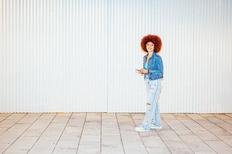 African descendent woman with vibrant afro hair, wearing a denim jacket and ripped jeans, stands smiling against a striped background, holding a smartphone