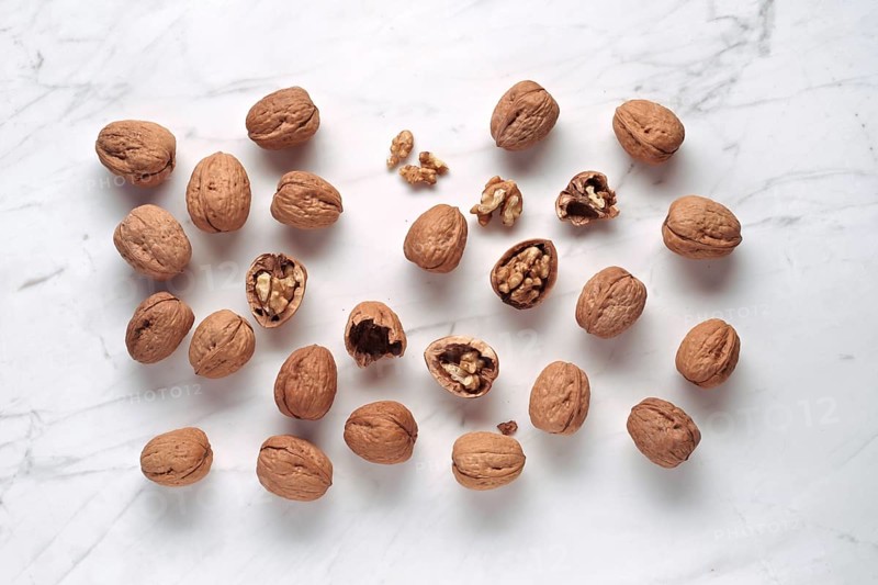 A collection of walnuts, some cracked open to reveal the kernels inside, scattered on a white marble surface. The nuts display their rich brown shells and intricate textures