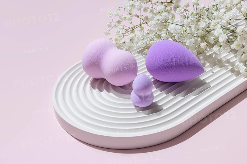 Various sizes and shapes of purple-toned makeup sponges expertly staged on a white ridged tray, accompanied by delicate white flowers on a pink background