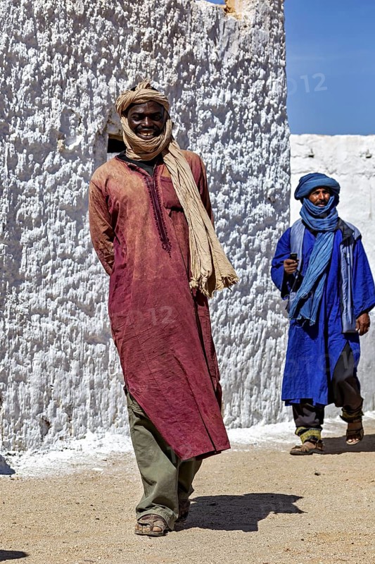 Two men in traditional Tuareg dress walk in front of a white plastered wall, local people in the Sahara in Algeria