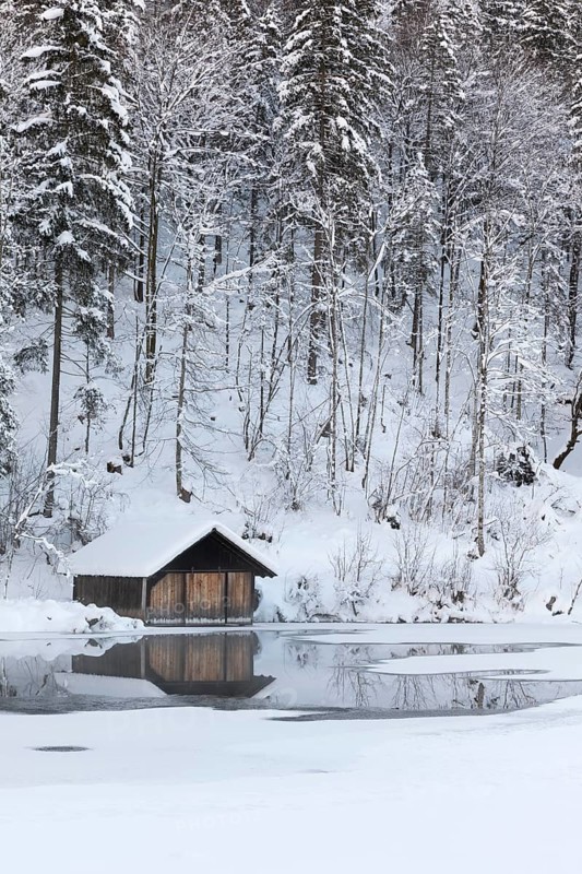 Snow covered boat shelter on Lake Almsee which is partly frozen and snow covered. Snow on the forest with gray sky