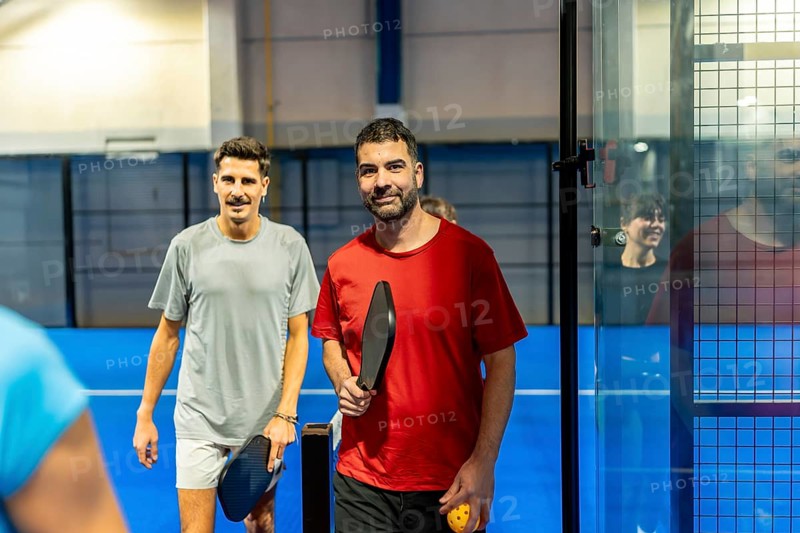 Two smiling men stand on a blue indoor pickleball court holding paddles and a ball, ready to play and enjoy a friendly, active game promoting fitness, fun, and social recreation